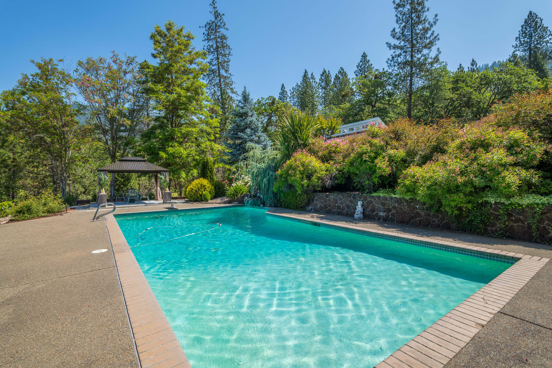 Rectangular in-ground pool with brick coping, a curtained gazebo at left, and a rock waterfall at the far end